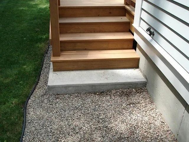 Wooden outdoor steps with a small concrete landing beside a gravel path and house siding.