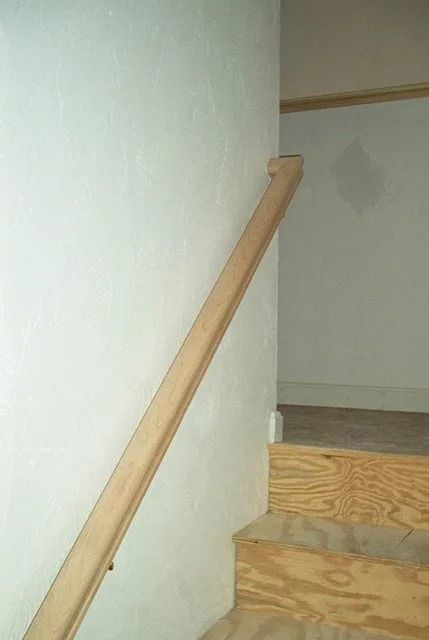 Wooden handrail beside a narrow indoor stairway with white walls and unfinished steps.
