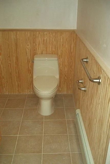 Small bathroom with a toilet, wall-mounted grab bars, and tan tile walls and floor.