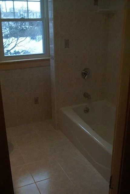 Dim bathroom with a bathtub, tiled floor, and a window letting in daylight.