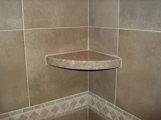 Corner shower shelf made of tan stone tile with a small decorative ledge.