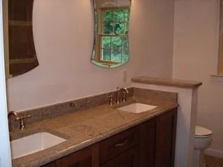 Bathroom vanity with double sinks, wood cabinets, tan countertop, and a toilet beside a small window.