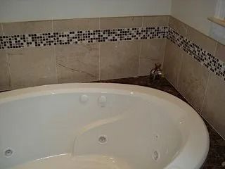 White corner bathtub in a tiled bathroom with mosaic trim and a faucet on the right