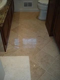 Bathroom floor with beige tile, a toilet at right, and a white bath mat in the foreground