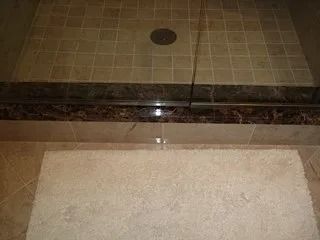 Bathroom shower with beige tile, dark trim, and a light bath mat in front.