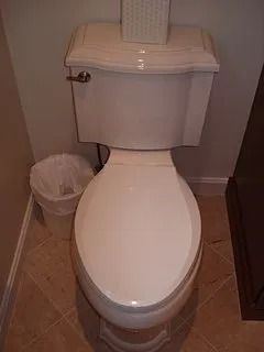 White toilet with closed lid in a small bathroom, with a trash can beside it.