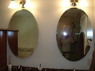 Two oval bathroom mirrors above sinks, lit by wall sconces, with a wooden cabinet on the left.