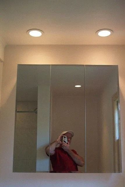Person taking a mirror selfie in a bathroom with two ceiling lights and mirrored cabinets.