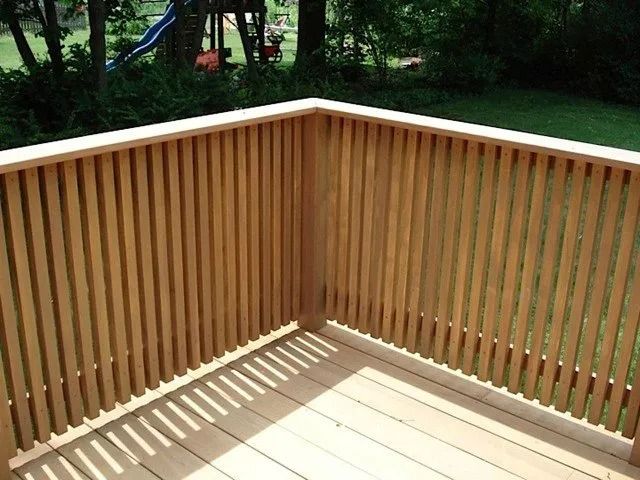 Wooden deck corner with vertical railings casting shadows on light floorboards, overlooking a green yard.