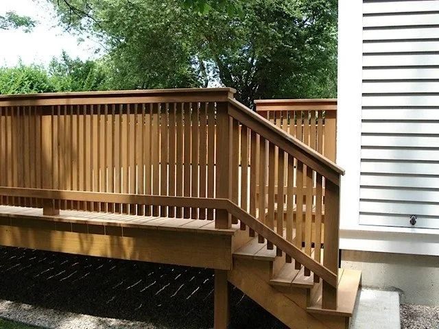 Wooden deck stairs with railing beside a white house, with trees in the background