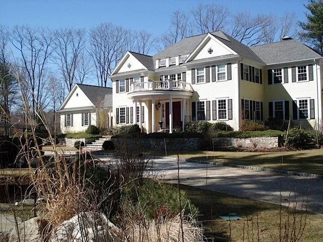 Large white colonial-style house with black shutters and a porch, viewed from a front yard on a sunny day