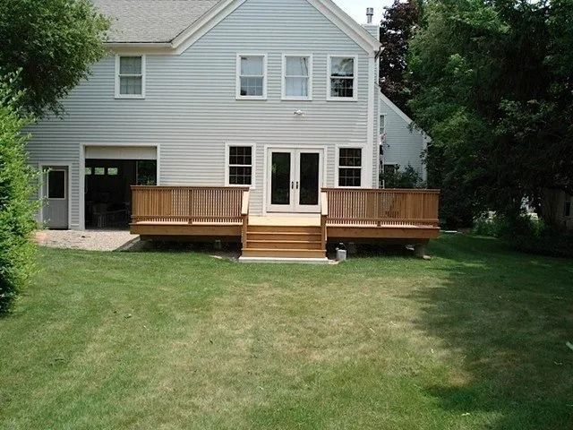 Backyard with a gray house and wooden deck, overlooking a grassy lawn and trees