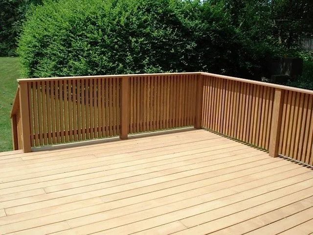 Wooden deck with tan floorboards and railing, surrounded by green bushes and trees