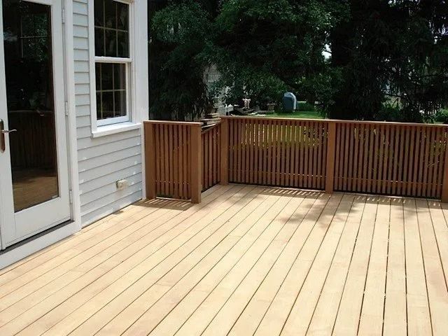 Wooden backyard deck beside a house, with sliding door, window, and railing overlooking trees.