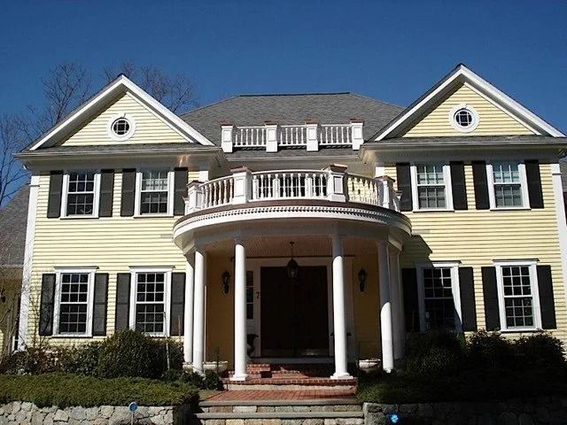 Large yellow colonial house with white columns, black shutters, and a covered front porch under a blue sky