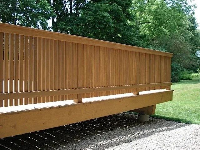 Wooden deck with vertical slat railing extending over gravel beside a grassy yard and trees