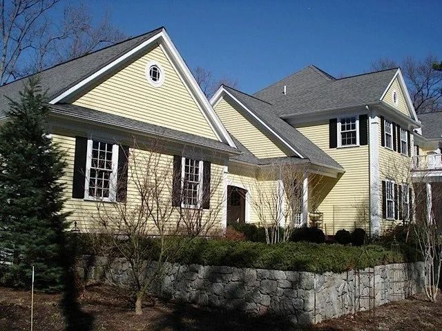 Yellow suburban house with gray roof, stone facade, and front porch behind a low stone wall.
