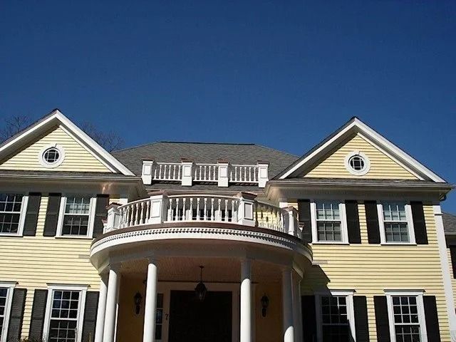 Large yellow colonial-style house with a white-columned porch and balcony against a blue sky