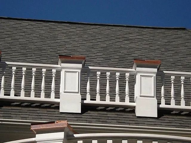 White balcony railing on a gray shingled roof with two dormer windows under a blue sky