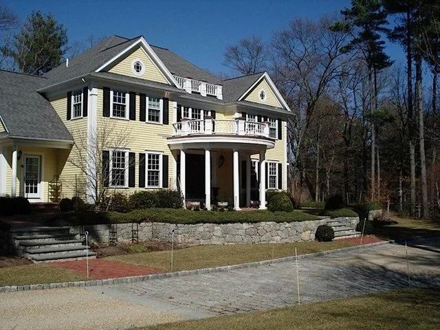 Large yellow two-story house with white columns, stone wall, and wooded yard under a clear blue sky
