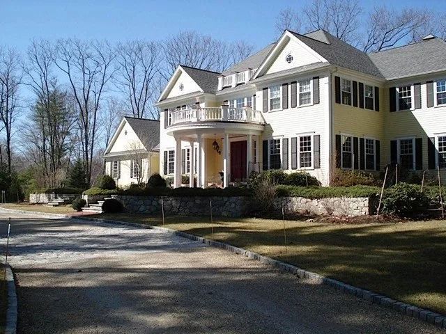 Large white two-story house with a curved driveway and front porch in a wooded suburban setting