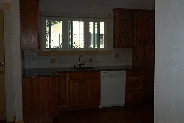 Dim kitchen with wooden cabinets, sink under a window, and a white dishwasher.