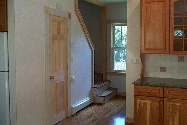 Small kitchen with wooden cabinets, tiled backsplash, and a stairway beside a window.