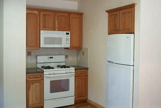 Small kitchen with wooden cabinets, white stove and microwave, and a white refrigerator against a beige wall.