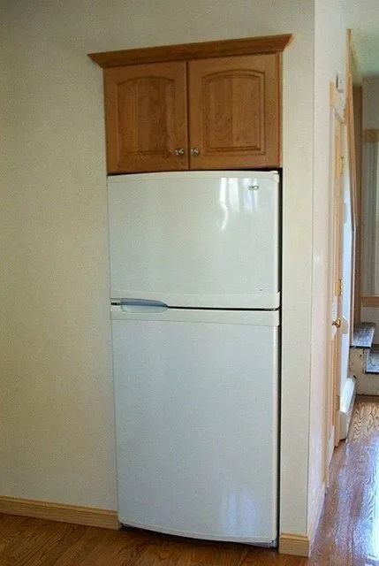 White refrigerator beneath wooden cabinets in a narrow kitchen hallway.