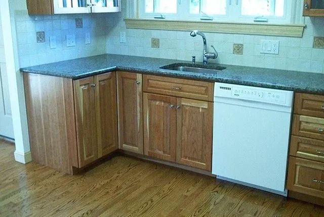 Small kitchen with wooden cabinets, dark countertop, white dishwasher, sink, and hardwood floor.