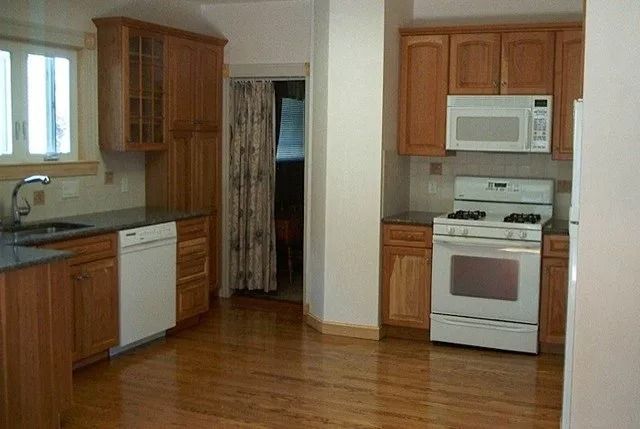 Empty kitchen with wooden cabinets, white appliances, and hardwood floors, viewed from the dining area