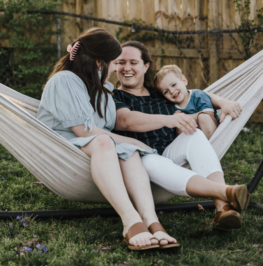 Three people sit in a white fabric hammock in a grassy outdoor area, laughing together.