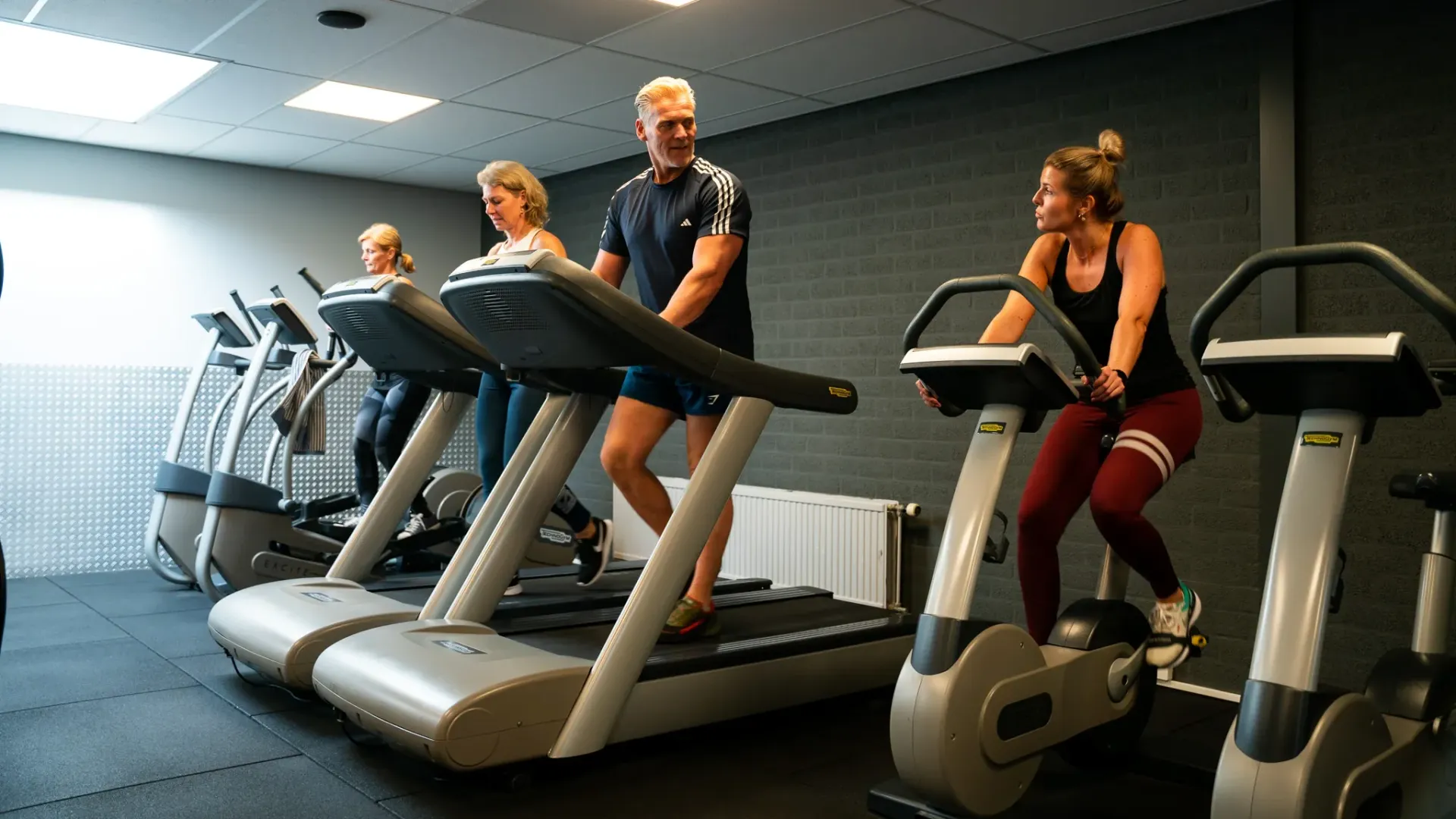 Een groep mensen rent op loopbanden, daarlangs fiets een vrouw op een hometrainer, daarlangs gebruikt een vrouw een elliptical trainer in een sportschool.
