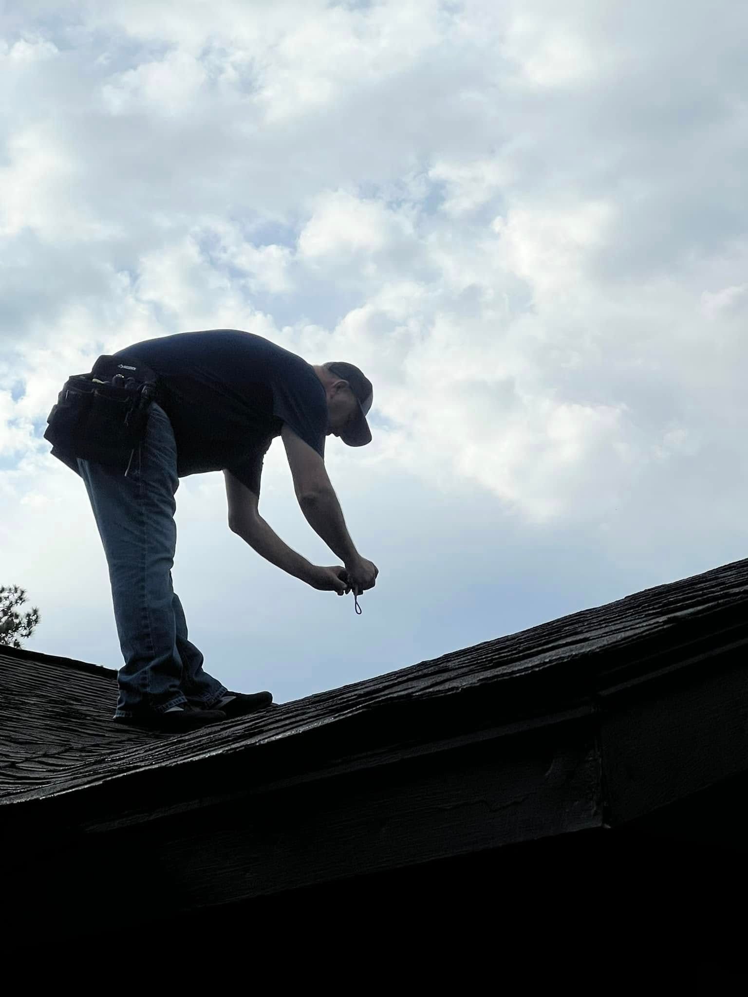 Person bending over on a rooftop, working on dark shingles under a cloudy sky