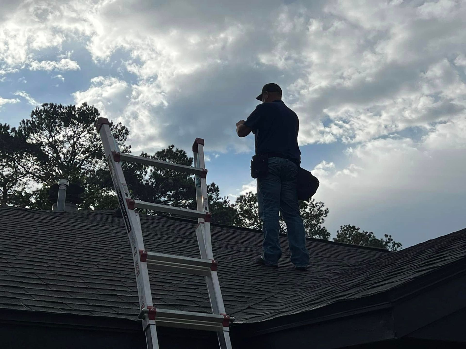Worker on a shingled roof beside a ladder, inspecting or repairing the roof under a cloudy sky.