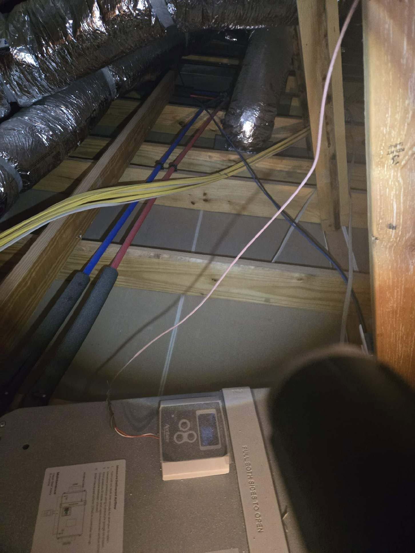 Attic with exposed ductwork, insulated pipes, and cables over wooden framing; HVAC unit in foreground