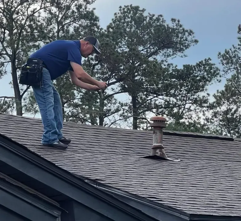 Worker inspecting roof shingles on a house with trees in the background