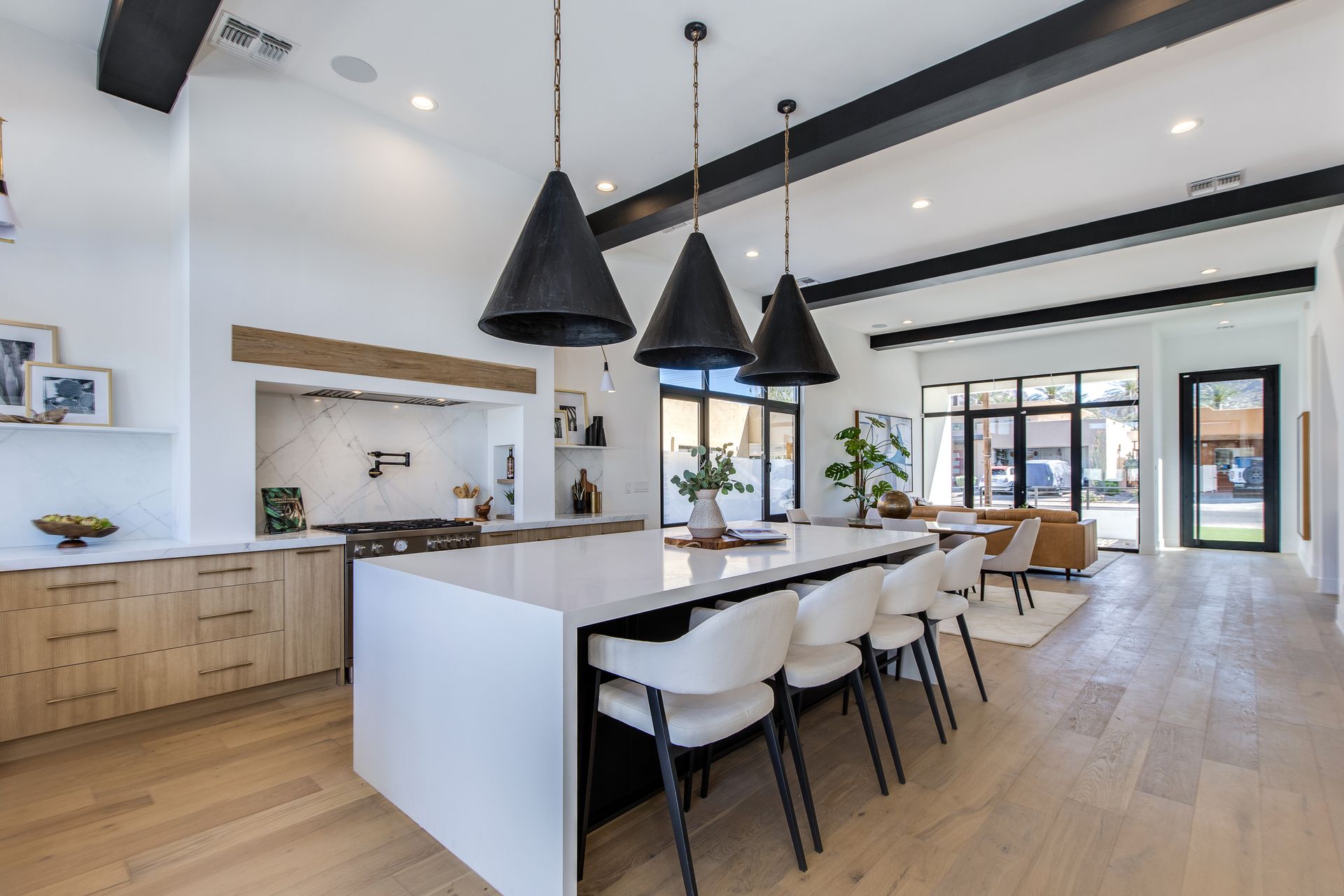 A modern, airy kitchen with light wood cabinets, a large white island with four stools, and three black hanging pendants.