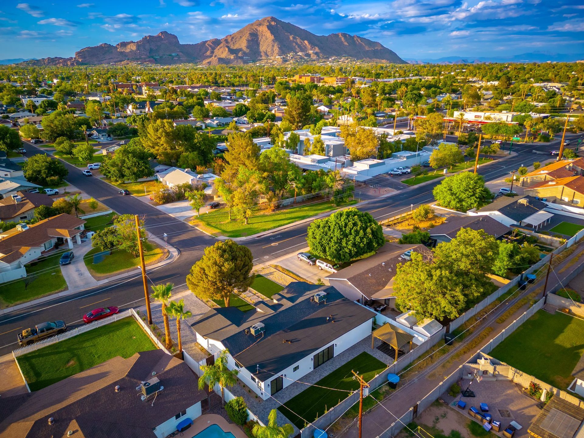 An aerial view of a suburban neighborhood with houses, green trees, and a mountain range in the distance under a blue sky.