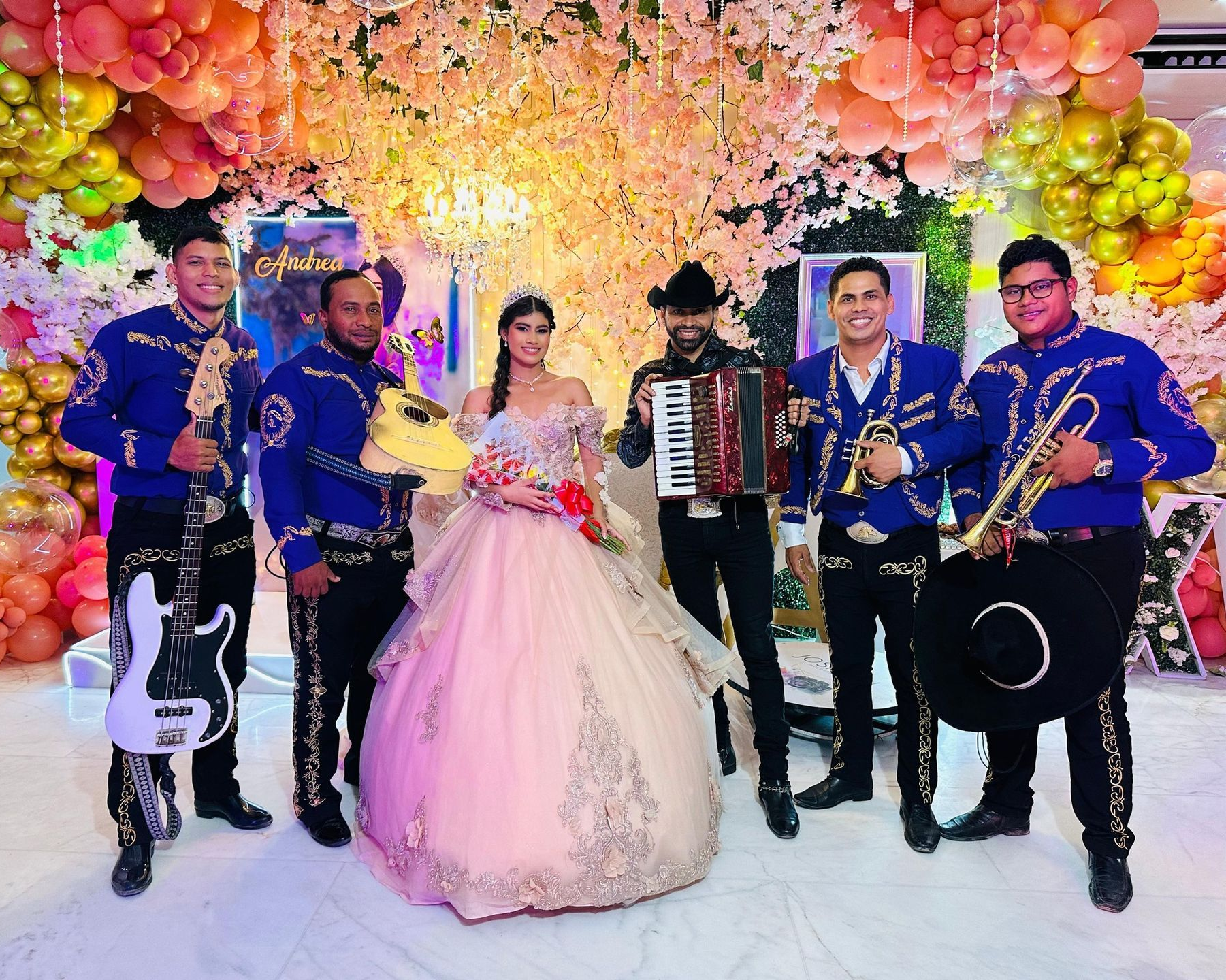 Una quinceañera posa con una banda de mariachis frente a un fondo floral con globos.