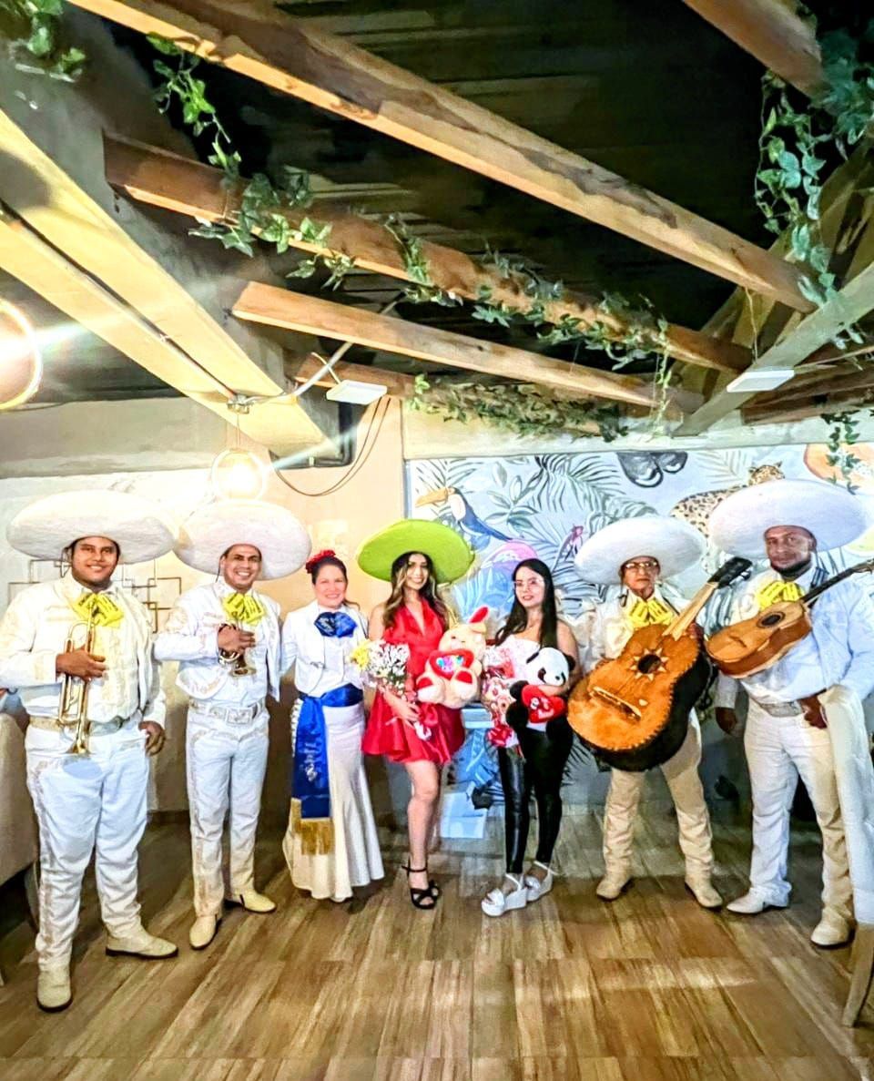 Banda de mariachis con dos mujeres en una sala con vigas de madera y vegetación.