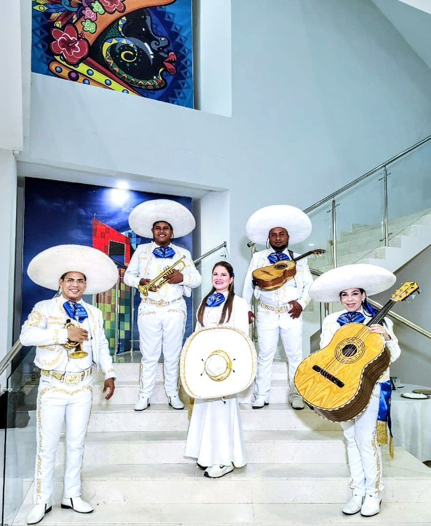 Banda de mariachis con trajes blancos en las escaleras, sosteniendo instrumentos, sonriendo y actuando.