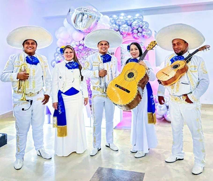 Banda de mariachis con trajes blancos y instrumentos, sonriendo en una habitación decorada con globos.