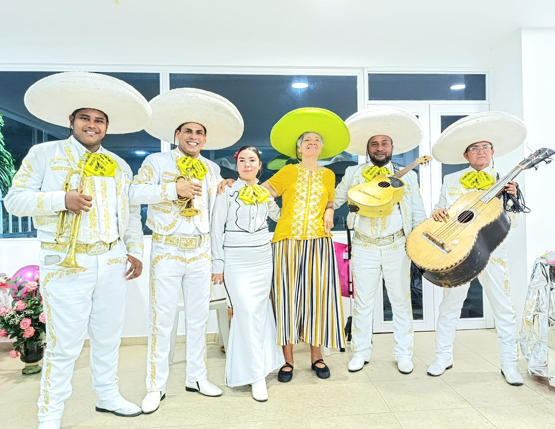 Banda de mariachis con trajes blancos y sombreros grandes, posando con una mujer con top y falda amarillas.