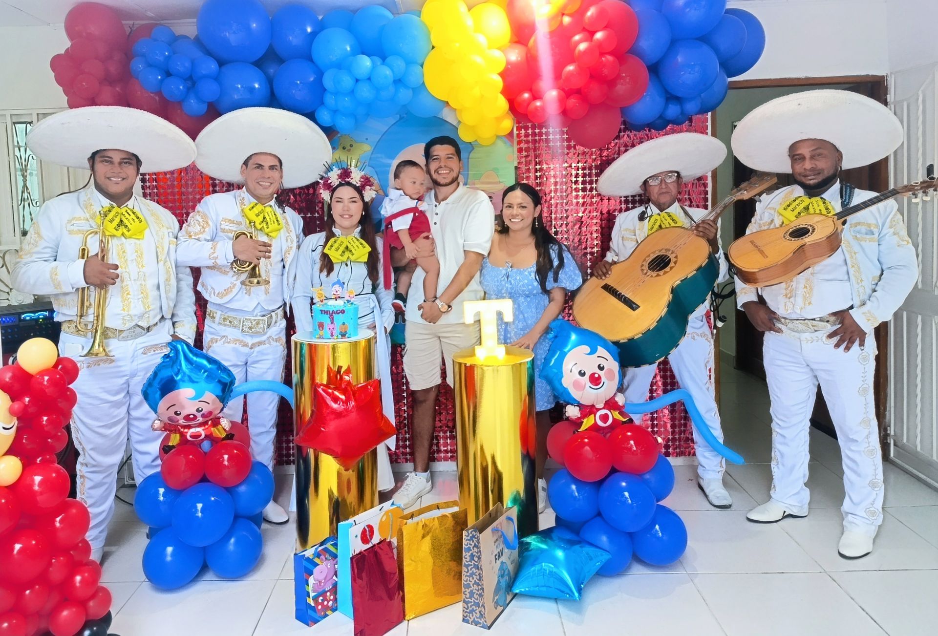 Banda de mariachis con familia en una fiesta de cumpleaños, globos azules y rojos, pastel, regalos.