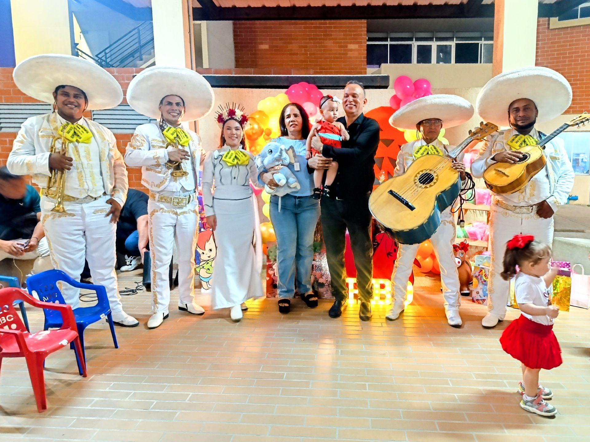 Mariachis con familia en una fiesta, con globos de fondo. Chica con falda roja.