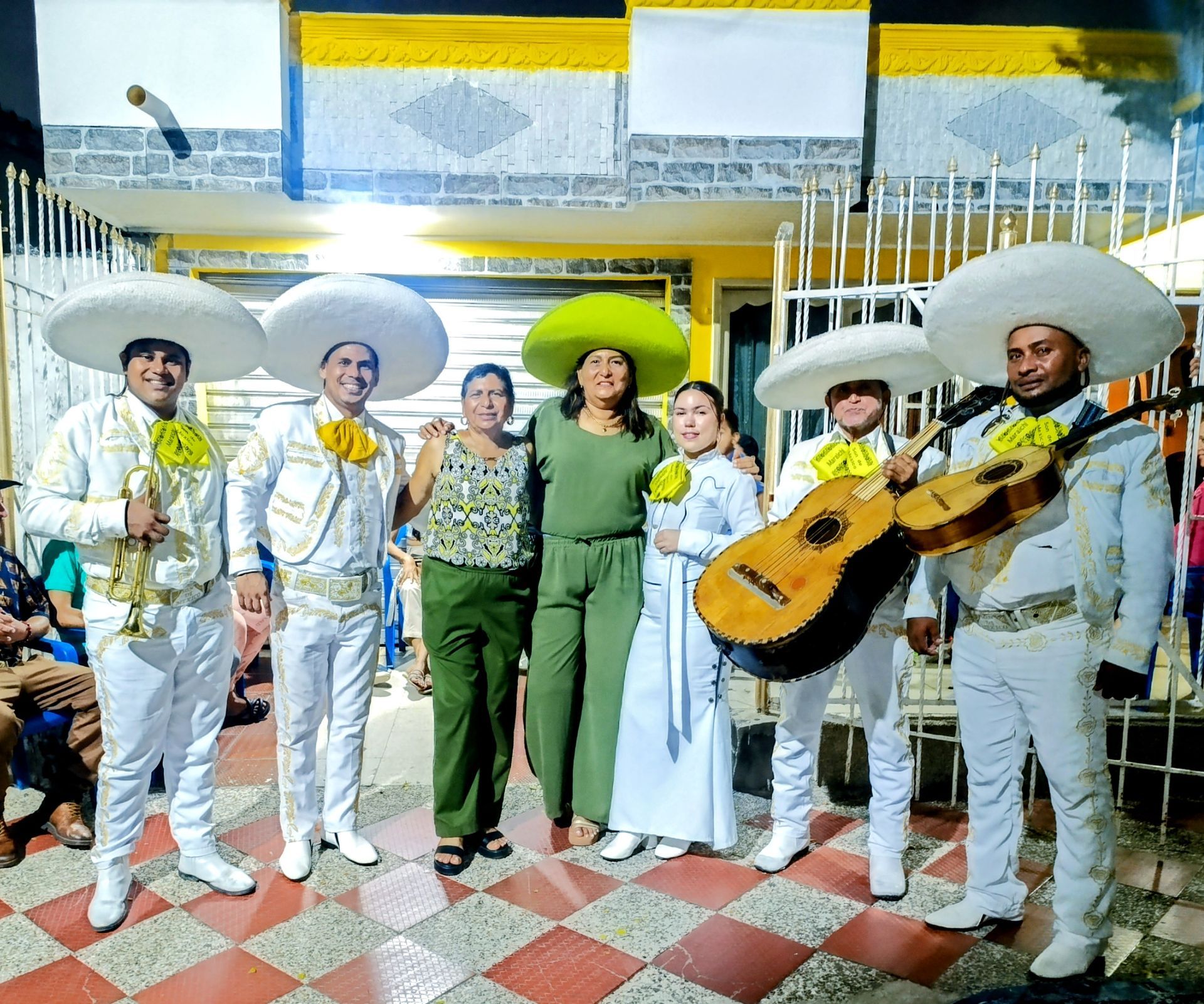 Banda de mariachis con trajes blancos y guitarra, posando con dos mujeres frente a un edificio.