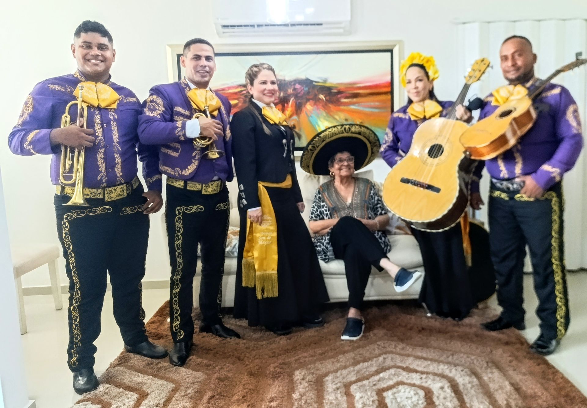 Banda de mariachis con mujer con sombrero, sonriendo, posando en una habitación con pintura.
