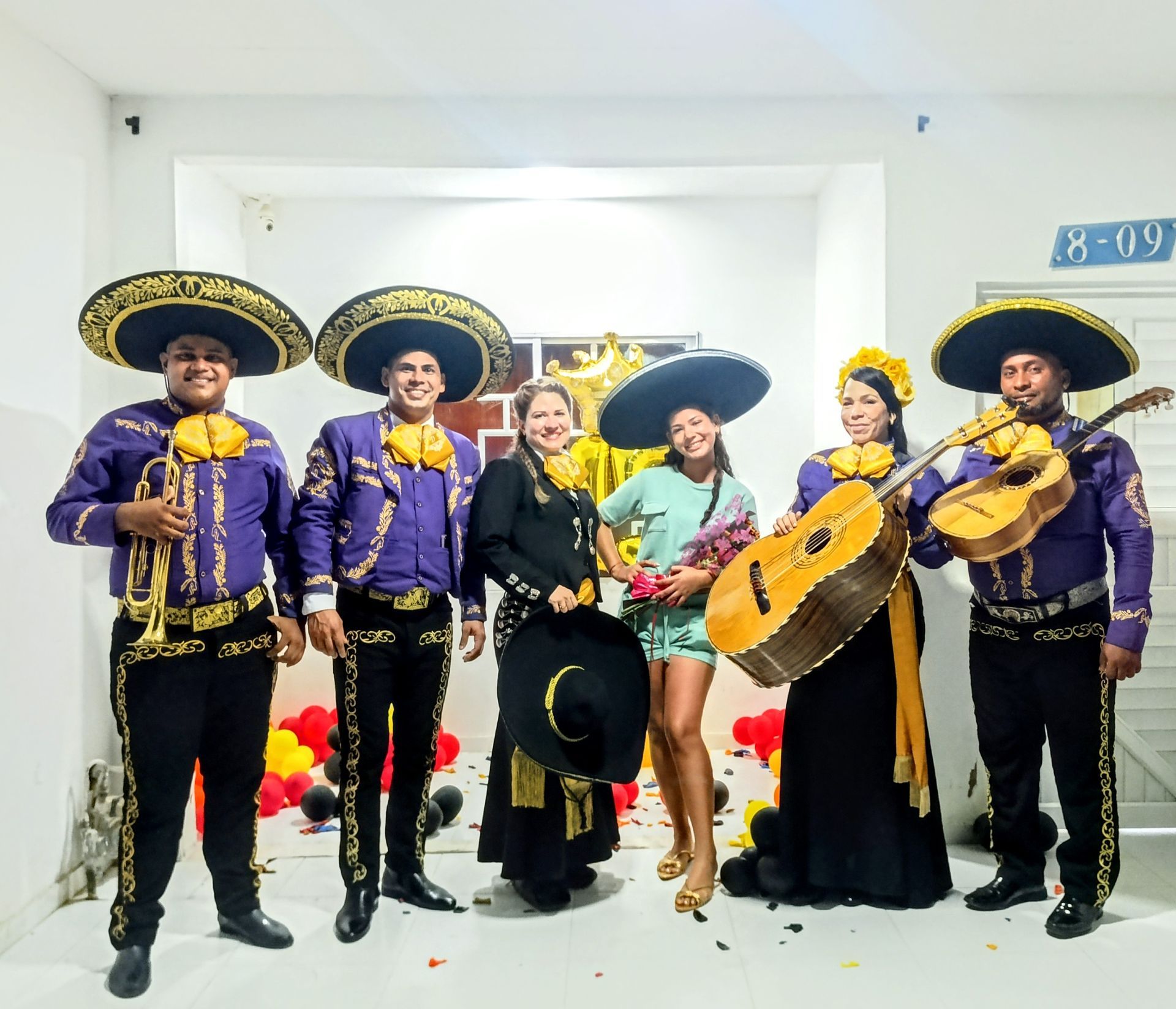 Banda de mariachis con dos mujeres, celebrando en el interior, atuendo morado y dorado.