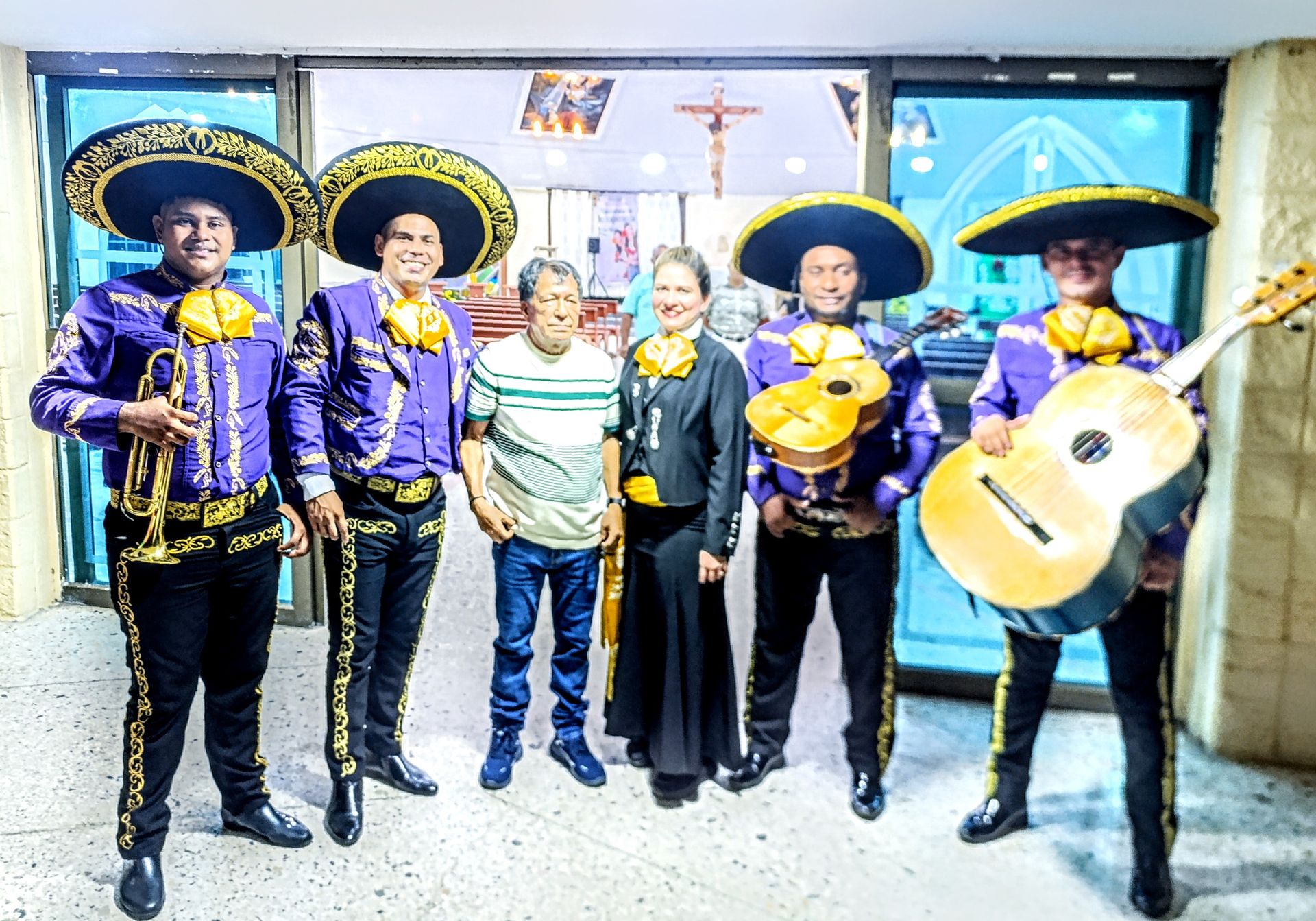 Banda de mariachis con trajes morados posa con dos personas frente a la entrada de la iglesia.